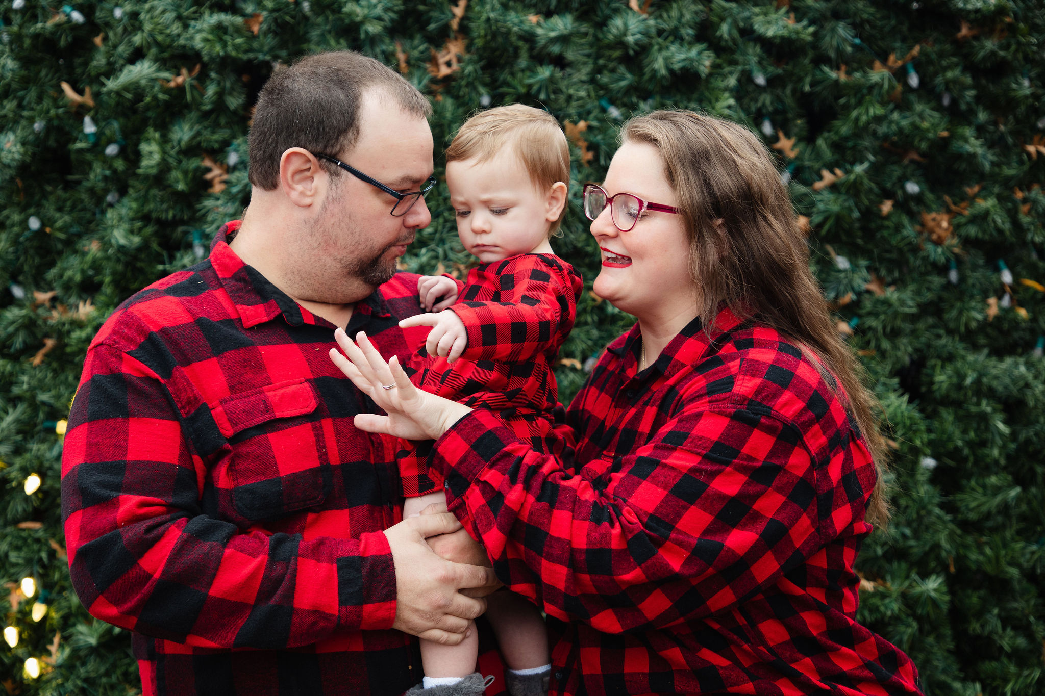 Parents sharing a tender moment with their toddler in front of a lit Christmas tree — candid family portrait by Karmil Studios