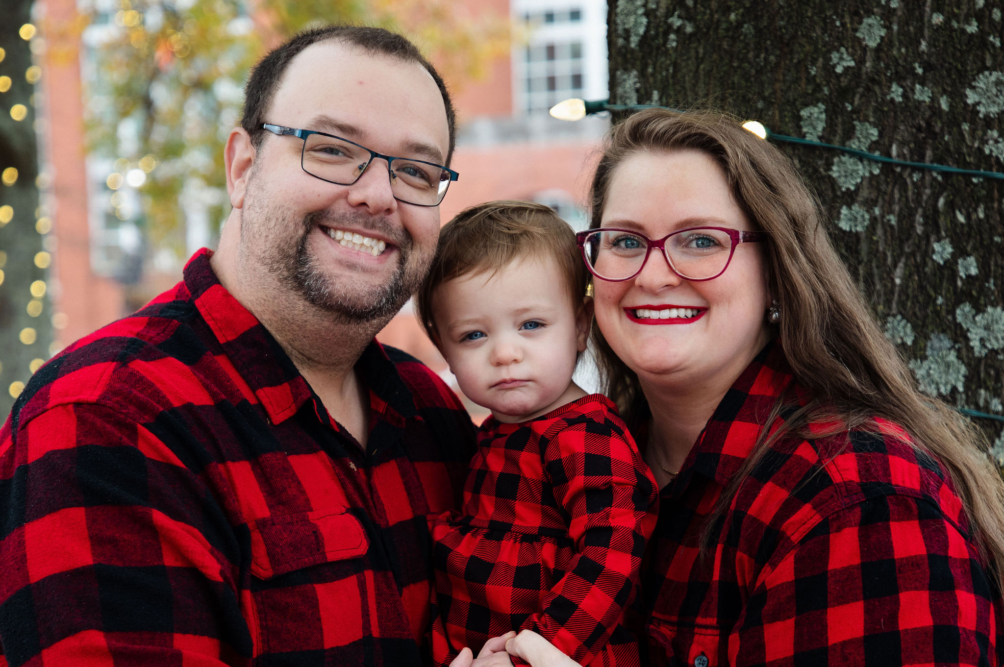 Close-up portrait of a family of three in matching red plaid with soft bokeh lights — holiday session by Karmil Studios