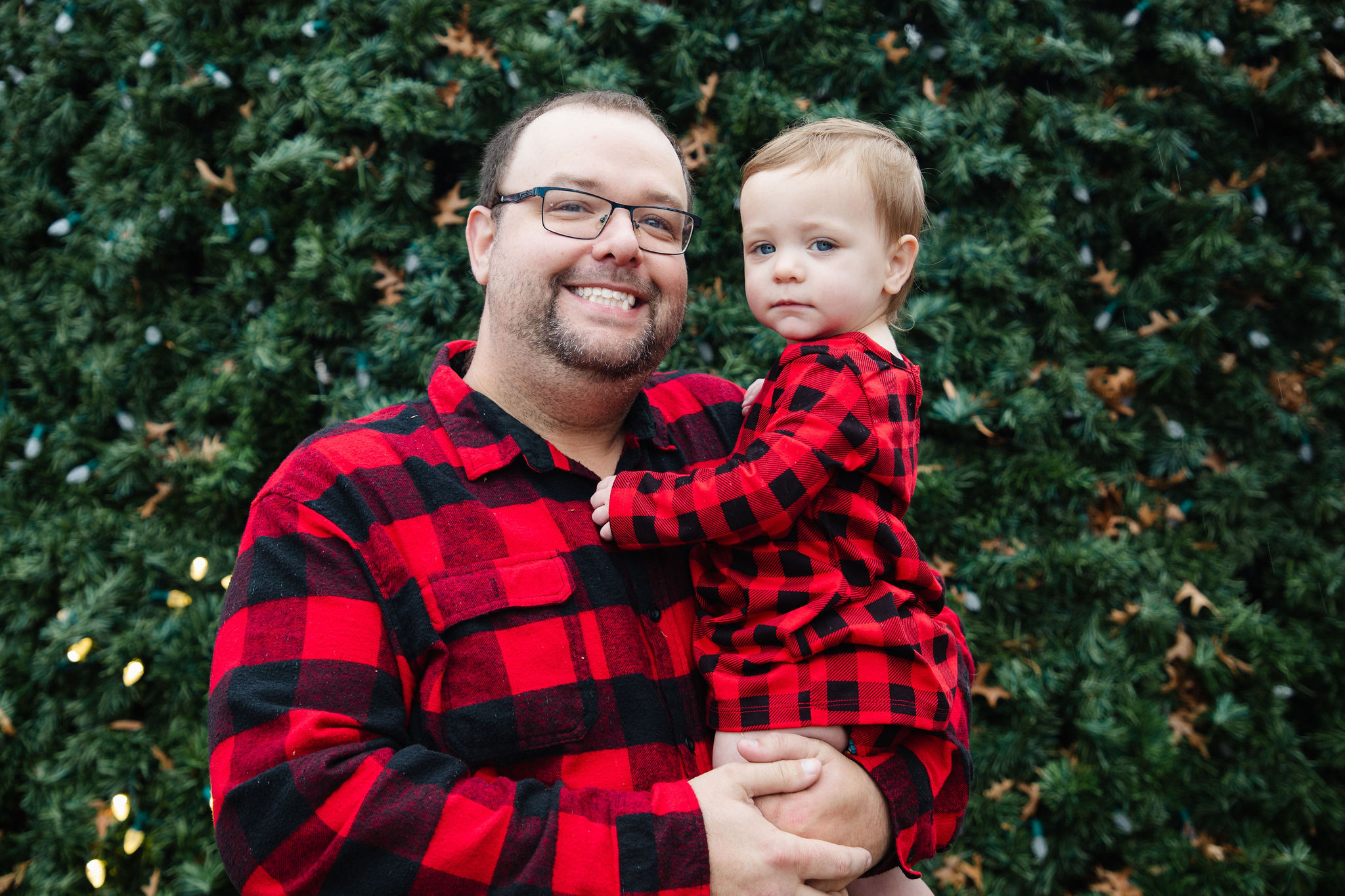Father holding his toddler in matching red buffalo plaid in front of a Christmas tree — family holiday portrait by Karmil Studios