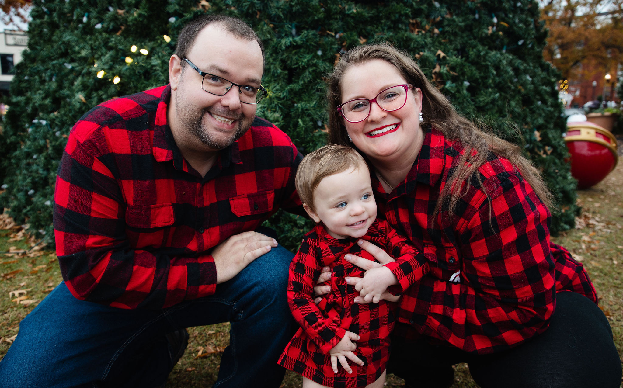 Family of three in matching red plaid kneeling in front of a large outdoor Christmas tree during a holiday portrait session by Karmil Studios