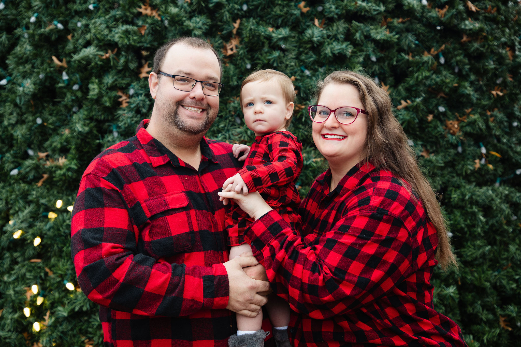 Family of three in matching red buffalo plaid smiling in front of a lush Christmas tree wall — holiday portrait by Karmil Studios