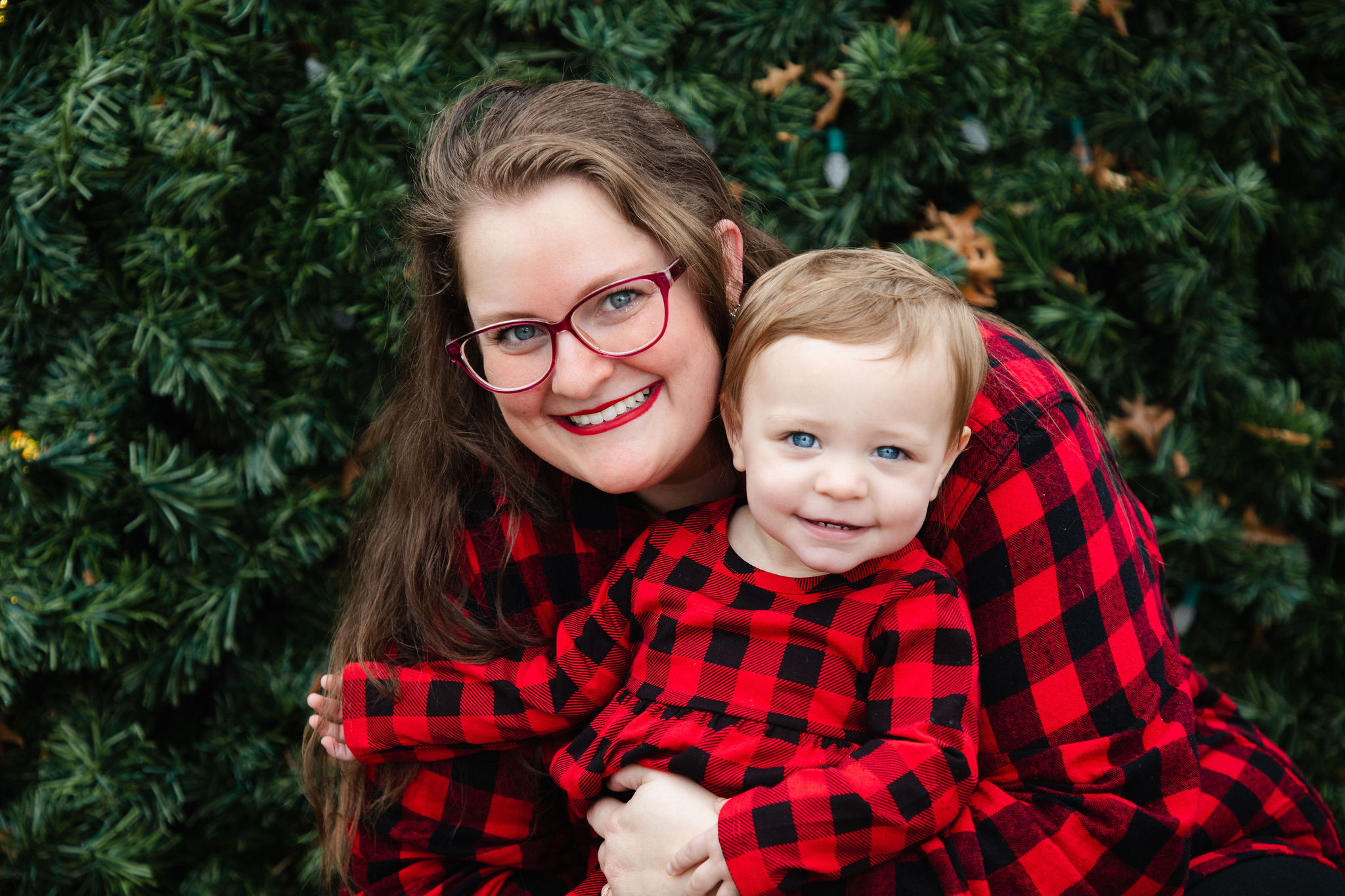 Mother and toddler smiling together in matching red plaid in front of a Christmas tree — holiday portrait by Karmil Studios