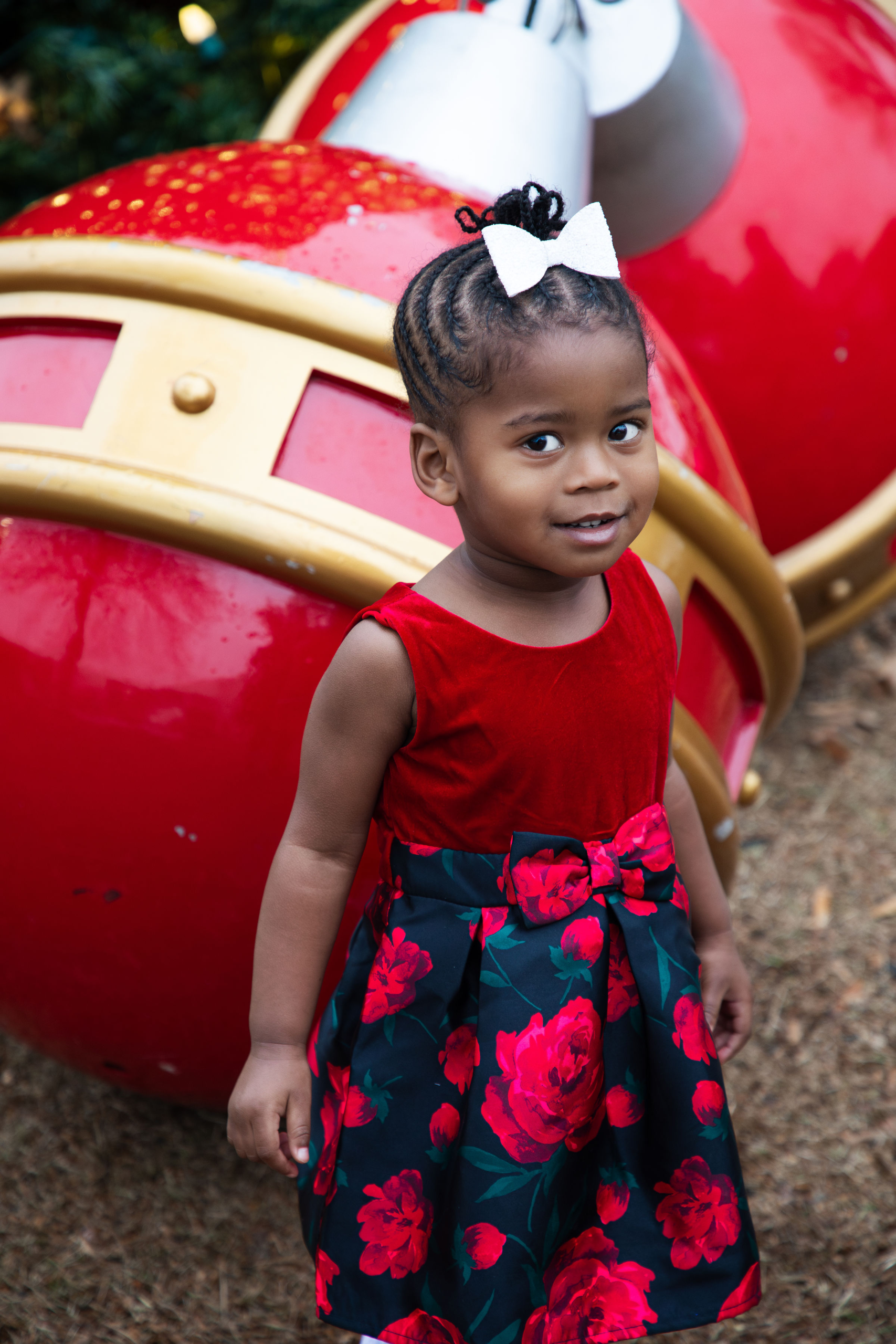Toddler girl in red floral holiday dress standing beside oversized Christmas ornament during holiday family photo session