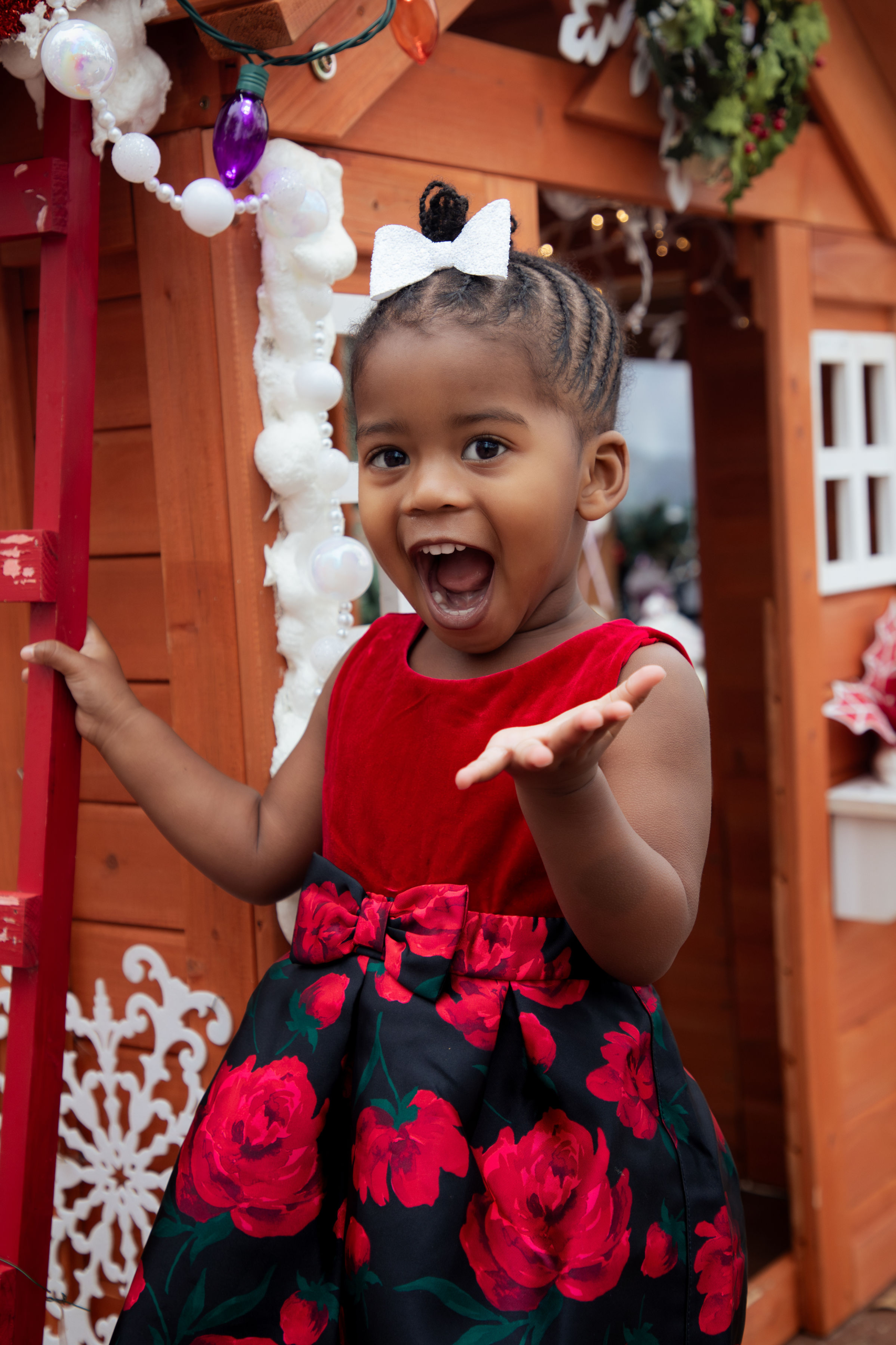 Toddler girl smiling and posing outside decorated Christmas playhouse during holiday portrait session