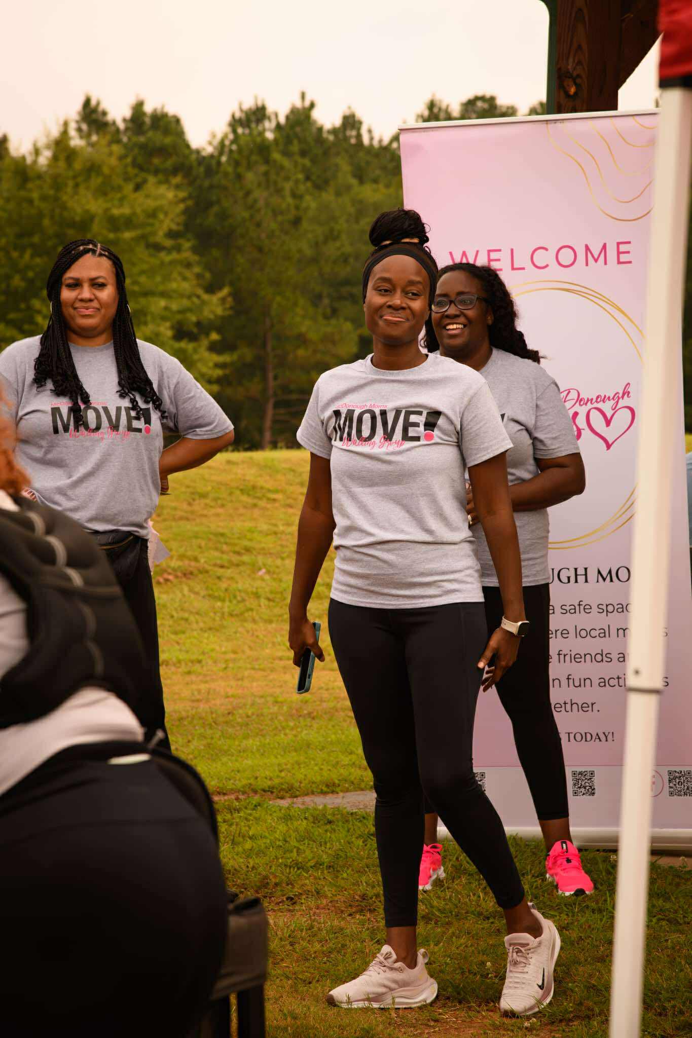 McDonough Moms MOVE walking group leaders welcoming attendees at a community wellness event beside a branded welcome banner.