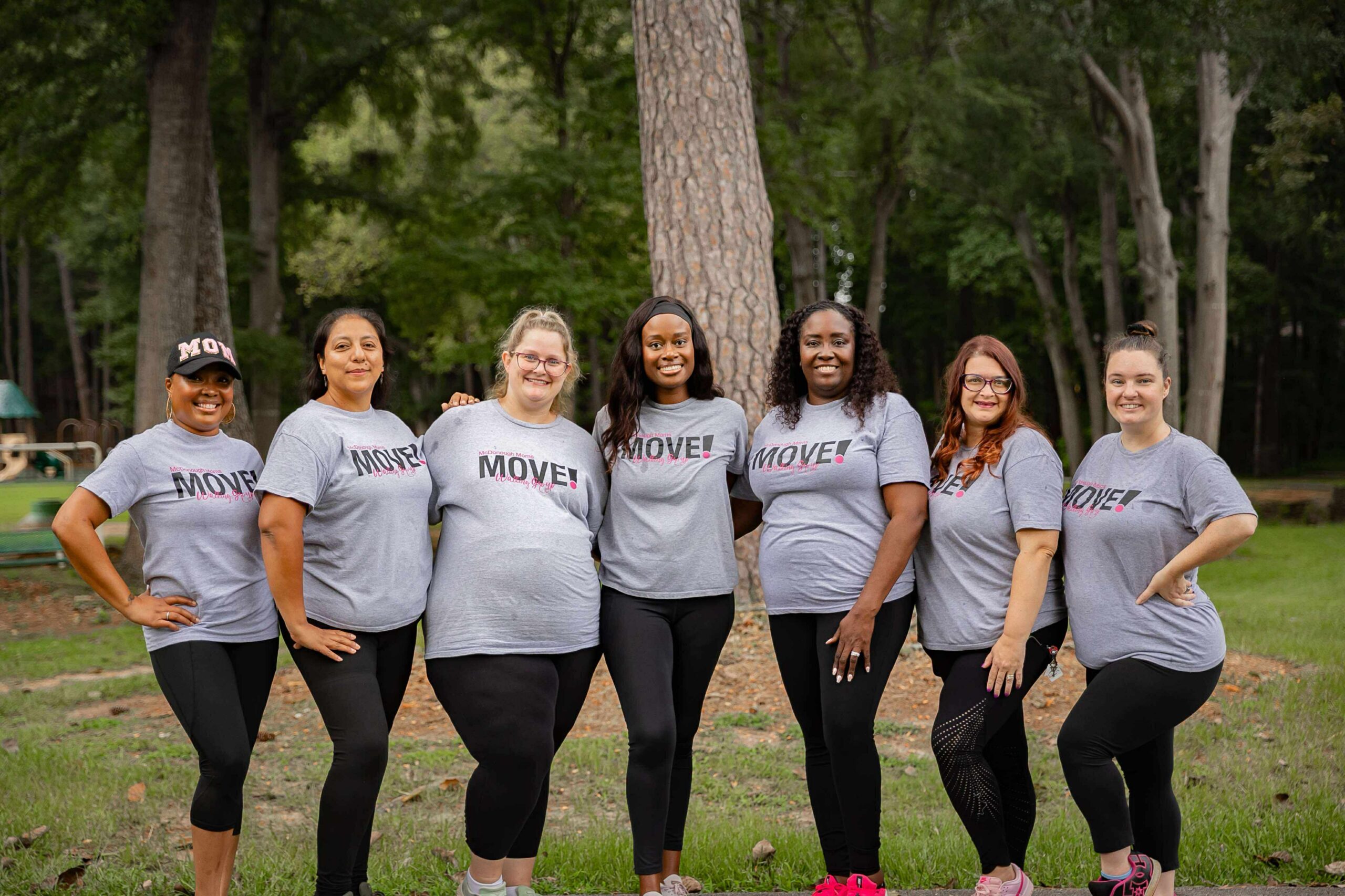 McDonough Moms MOVE walking group leaders posing together in matching gray MOVE shirts during a branded leadership photoshoot at a park.